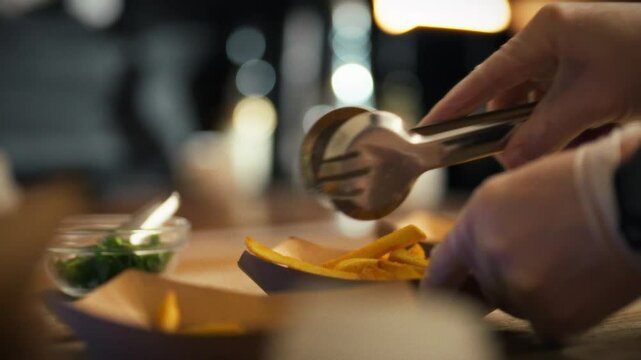 A young experienced fast food cook prepares food for delivery to customers. He puts freshly fried potatoes into paper dishes with metal tongs. The process of preparing orders for fast food customers
