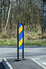 A vertical traffic sign featuring bold blue and yellow stripes stands on the roadside. Surrounding lush greenery suggests a spring setting, with trees visible nearby