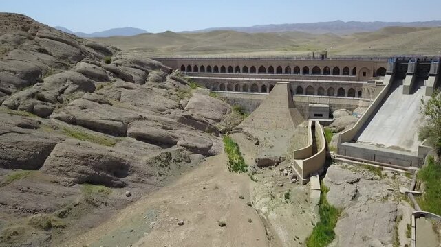 Drone shot directly above the dam capturing the greenish water in the upper reservoir and the cascading slope of the dam wall with multiple arched levels, offering contrast between nature and engineer