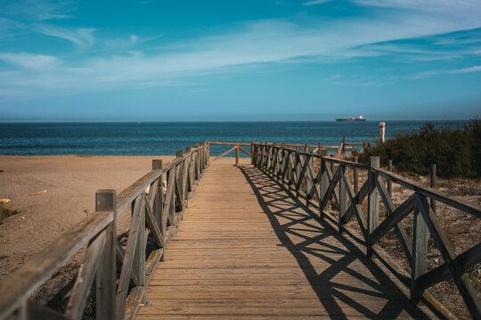 A scenic wooden pathway with railings leading to a serene beach and ocean view. The calm blue water and distant ship create a peaceful, relaxing atmosphere under a clear sky. High quality photo. 