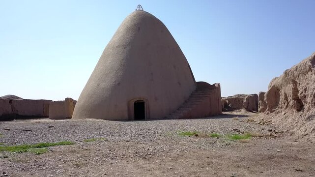 Drone flight above a traditional Persian yakhchal with a large conical dome and thick adobe walls in khorasan iran, surrounded by ruins of a desert village and green farmlands in the distance