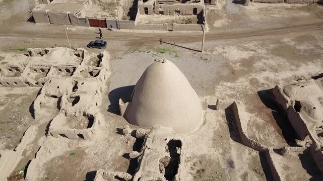 Drone flight above a traditional Persian yakhchal with a large conical dome and thick adobe walls in khorasan iran, surrounded by ruins of a desert village and green farmlands in the distance