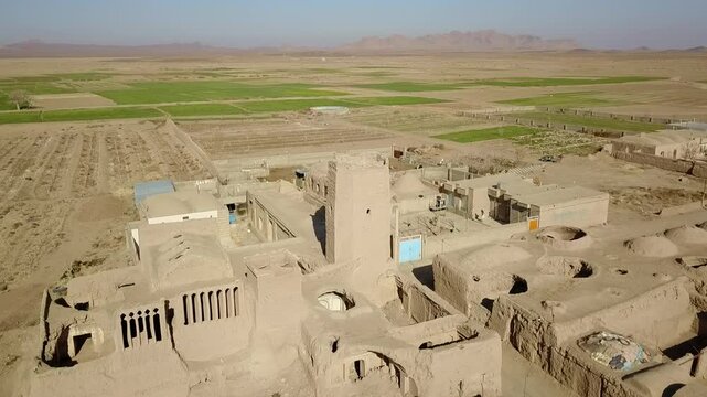 Aerial perspective of a ruined fortress and windcatcher structures surrounded by desert farms and scattered village homes. The scene depicts a blend of Persian environmental architecture, ancient