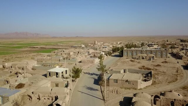 Aerial perspective of a ruined fortress and windcatcher structures surrounded by desert farms and scattered village homes. The scene depicts a blend of Persian environmental architecture, ancient