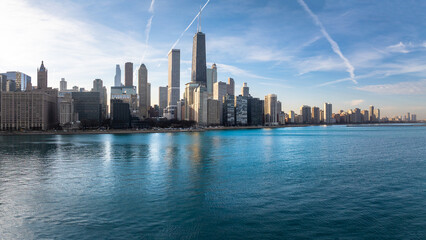 Fototapeta premium A stunning aerial view of the Chicago skyline along the Lake Michigan waterfront on a clear, sunny day. Skyscrapers rise above the shimmering blue water, with crisp contrails streaking across the vibr