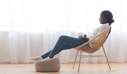 Relax at home. African american girl resting in wicker armchair and drinking coffee against window,...