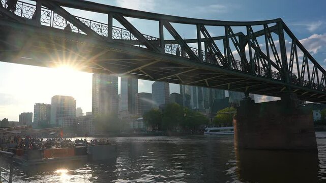 View of Eiserner Steg over the Main River in Frankfurt, with sunlight through ironwork, a boat in motion, and the modern skyline in the background.