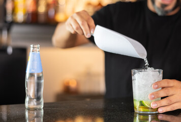 Unrecognizable male bartender prepares cocktail mojito in cafe, pours syrup into glass with ice. Glass is filled to top with crumb of ice, contains pieces of lime.