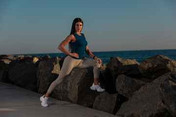 woman performs stretching exercise balancing on rocky shoreline as sun sets in background. ocean...