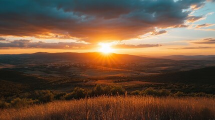 Sunset above landscape. Sun shining brightly. Sky with dark clouds. Golden light on grass and trees. Warm colors glow over the distant hills.