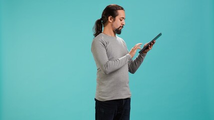 Portrait of upbeat person mindlessly scrolling on tablet screen, isolated over studio background. Cheerful man passing time on social media on portable device, enjoying leisure time, camera A © DC Studio