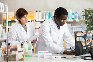 Fototapeta premium Two pharmacist employee using computer, reads service protocol update, finds out changes, and becomes familiar with work regulations. Woman and African man pharmacists at PC.
