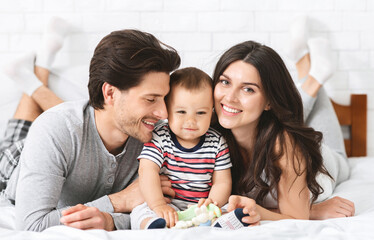 Enjoying parenthood. Happy young parents cuddling with adorable baby son on bed, free space