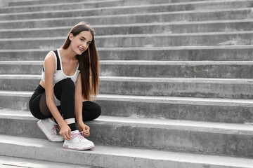 Obraz premium Young woman in sportswear and stylish sneakers tying shoe laces on stairs outdoors
