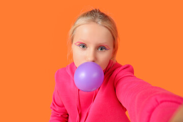 Cute trendy girl blowing bubble gum and taking selfie on orange background