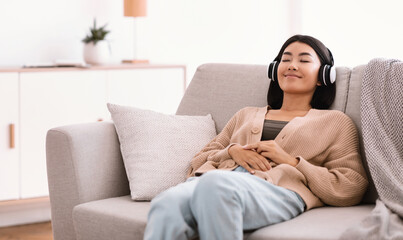 Obraz premium Rest And Relax Concept. Portrait of calm asian woman lying on the couch, listening to music, audio book or podcast, enjoying meditation for sleep and peaceful mind in wireless headset at home