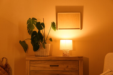 Interior of night living room with glowing lamp and houseplant on commode near yellow wall