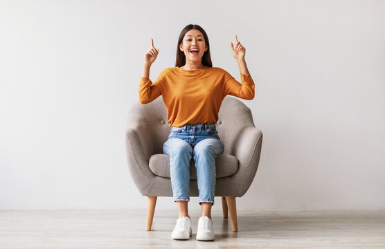 Excited young Asian woman sitting in armchair, pointing two fingers up, offering space for your advertisement against white studio wall, full length. Best promotion, cool discount, huge sale concept