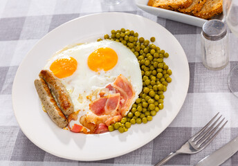 Plate contains scrambled eggs with slices of bacon, homemade sausages, complemented by vegetable side dish green peas. Sliced bread, plate of food, cutlery and glasses placed on checkered tablecloth