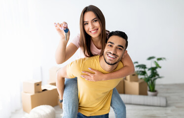 Cheerful Caucasian lady getting piggyback ride from her Arab boyfriend, showing house key indoors. Lovely multinational couple posing in new apartment on moving day