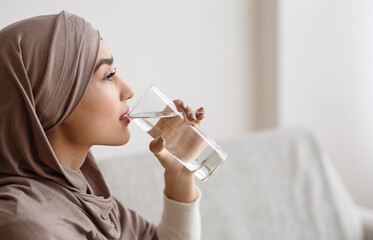 Thirsty islamic woman in headscarf drinking mineral water from glass at home, selective focus,...