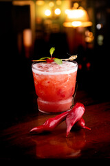 Spicy red cocktail with peppers and mint on angled pub table with blurred bar background