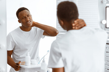 Fototapeta premium African Man Touching Aching Neck Suffering From Pain In The Morning, Standing Near Mirror In Bathroom At Home. Male Health Problems, Cervicalgia, Poor Posture And Headache Issue. Selective Focus