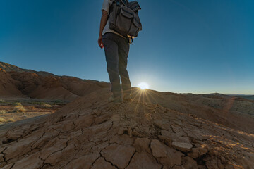 The traveler stands on a hill during the golden hour, when the sun is almost below the horizon. A man with a backpack, a photo from the back, at sunset.
