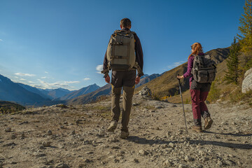 Two travelers in bright clothes with backpacks on their backs are walking along a mountain road to a slope with a magnificent view of a valley with a river and a forest.