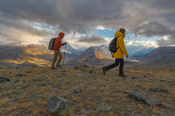 Travelers in bright clothes walk along the mountainside during the golden hour, overlooking the snowy mountains and the lake in the distance. Tourists with backpacks on their backs during sunset with 