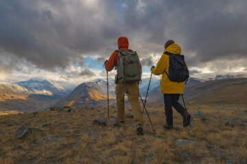 Urist travelers in bright clothes stand on a hill during the golden hour, in front of them there is a view of snowy mountains and valleys with lakes.