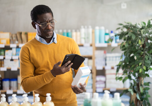 Focused middle aged african american man in casual ochre sweater and glasses scanning qr code on body lotion package(bottle, tube) with phone, browsing product information online in pharmacy..