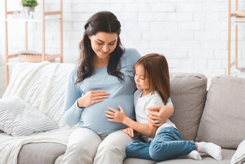 Happy pregnant mother and preschool daughter sitting on sofa at home, having conversation, copy...