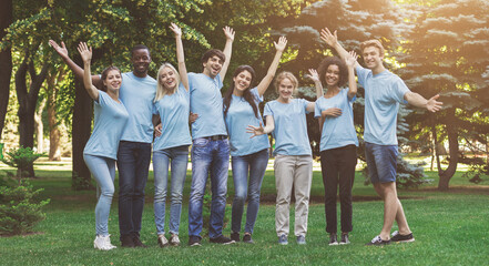 Happy millennial volunteers greeting you, waving hands and cuddling together at park, free space