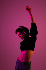 Cheerful young woman in wireless headphones feeling the rhythm, dancing with closed eyes in neon lights, against a red studio background.