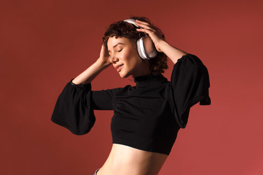 Young woman enjoying music with wireless headphones, smiling with eyes closed, wearing a black top, standing against a warm red background.