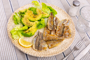 Piece of baked fish bank cod with side dish of fresh vegetables is served on plate. Fish is guaranteed with salad of spinach leaves and avocado slices, complemented by glass and cutlery.