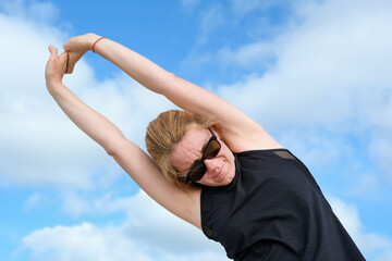 Woman stretching arms overhead outdoors under bright sky, enjoying fitness and wellness routine, focusing on flexibility, strength, and energy in a natural environment