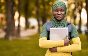 Fototapeta premium Education For Muslim Women. Smiling black student lady in hijab standing outdoors with workbooks, carrying backpack and posing to camera, islamic femle having free time at campus park, copy space