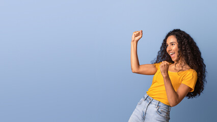 Euphoric brunette woman emotionally celebrating success, raising fists and exclaiming with excitement, lucky young lady yelling with joy, standing over purple background in studio, panorama