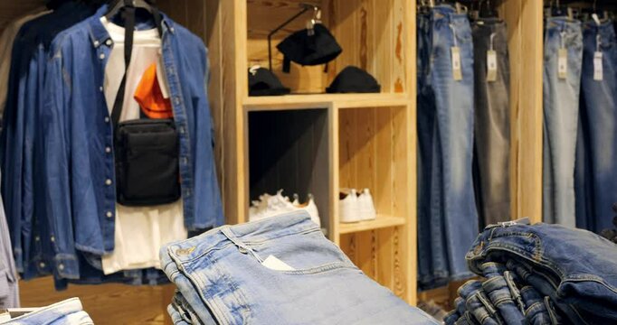 Stacks of folded jeans sit on a wooden display table, with shirts and accessories hanging on racks in the background, creating a visually appealing arrangement in a clothing store