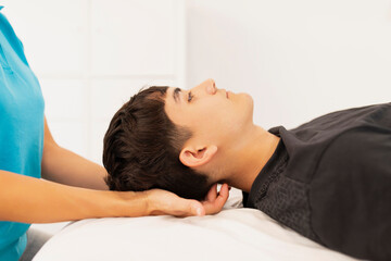 A young man, lying on a stretcher, receives a massage in the neck, cervical area, by a physiotherapist