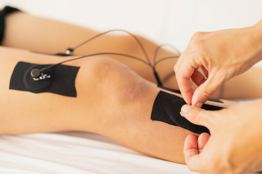 A physiotherapist places the electrodes on the leg of a young patient during an electrostimulation treatment, due to a sportive injury on the knee
