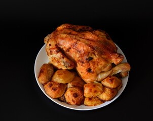 White ceramic plate with new potatoes and whole baked chicken on black background.