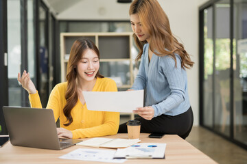 Two business women using laptop and tablet sitting and standing together to review documents in the office with her female friend.