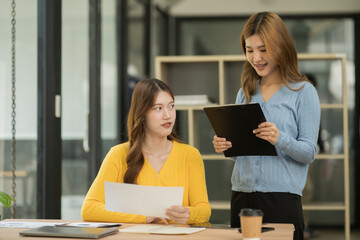 Two business women using laptop and tablet sitting and standing together to review documents in the office with her female friend.