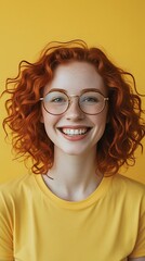 Obraz premium Cheerful young woman with red curly hair and glasses smiling brightly against a vibrant yellow background, radiating positivity, confidence, and natural beauty.