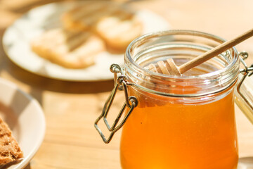 A jar of honey sits in a warm, sunlit kitchen with a wooden dipper resting inside. Next to it are slices of fresh bread on a plate, creating an inviting atmosphere.