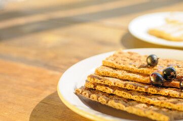 Crunchy crackers are arranged on a rustic wooden table, topped with fresh blueberries. Natural sunlight enhances the warm and inviting setting.