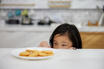child girl hiding and looking butter cookies or biscuits on dish from under the table in the kitchen